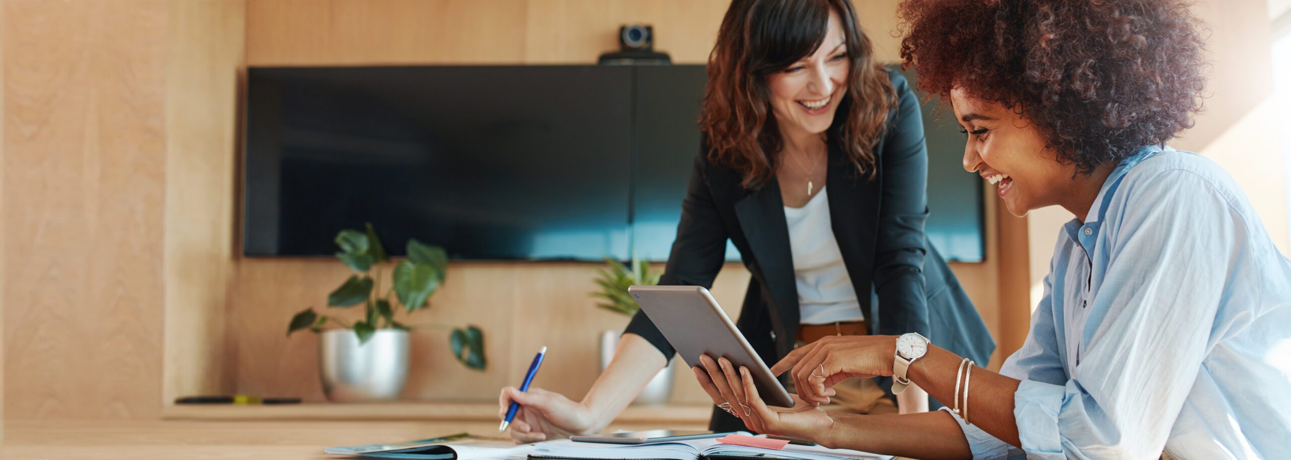 Two women in an office setting smiling and discussing something on a tablet. One is sitting, and the other is standing, holding a pen. A television and potted plant in the background.